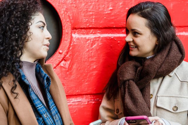 two women in their twenties sitting in front of a red wall apparenty talking